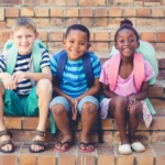 Three elementary school kids with backpacks sit on the steps of a school. Robuck homes builds in communities that have some of the best schools in North Carolina.